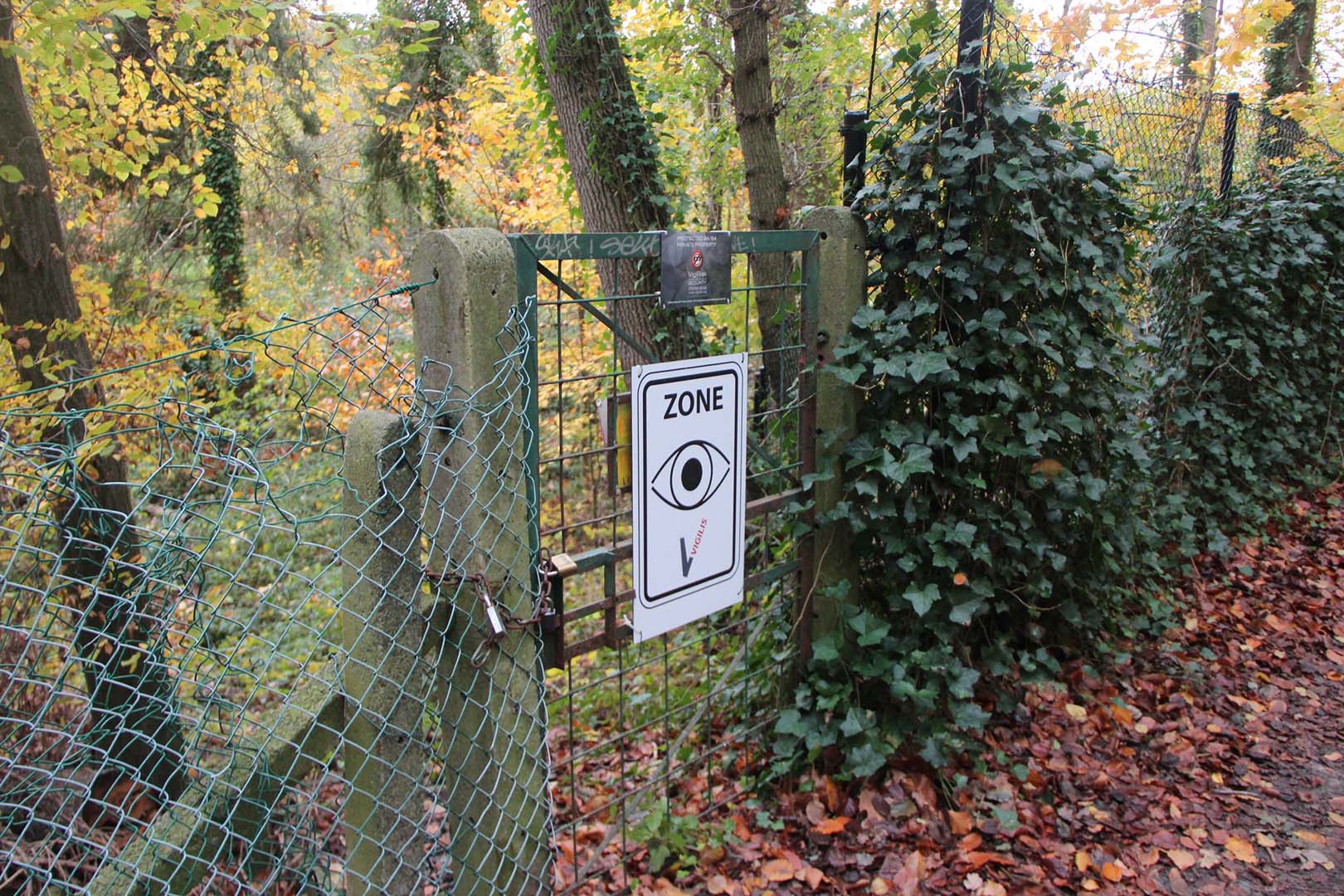 Chemin piéton entouré d'arbres, ancienne entrée du campus condamnée, un panneau sur l'entrée prévient de la présence d'une société de sécurité qui surveille le site